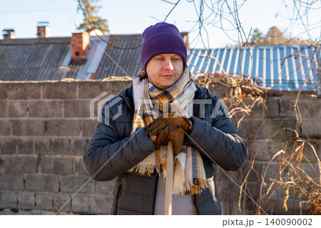 Gardener man wearing warm hat and scarf holding wooden rake in backyard. Rural worker engaged in autumn garden maintenance. Agricultural and seasonal yard care lifestyle concept. 140090002