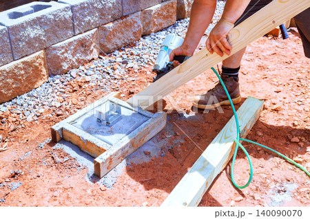 Construction worker secures wooden board to concrete base using nail gun during building project outdoors. 140090070