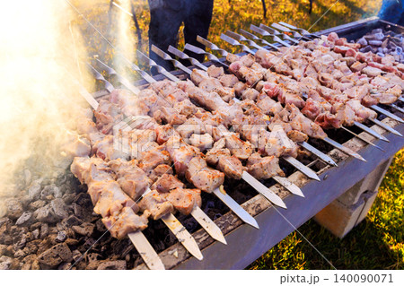People cook skewers of meat over hot coals at barbecue outside on sunny afternoon. People cook skewers of meat over hot coals at barbecue outside on sunny afternoon. 140090071