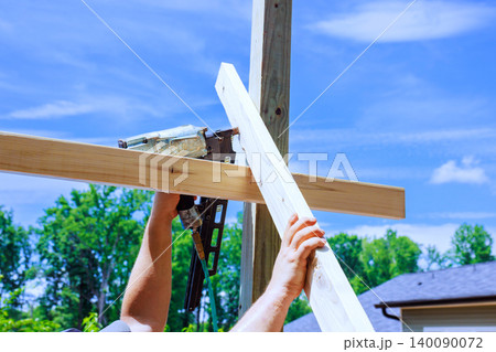 Worker is working on construction project by using nail gun to secure wooden beams against post in yard. Worker is working on construction project by using nail gun to secure wooden beams against post in yard. 140090072