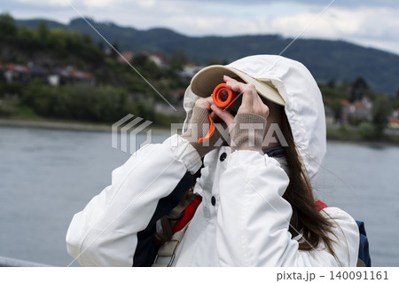 Tourist looking through a small orange telescope at a river landscape, emphasizing sightseeing and scenic viewpoints. Tourist looking through a small orange telescope at a river landscape, emphasizing sightseeing and scenic viewpoints. 140091161