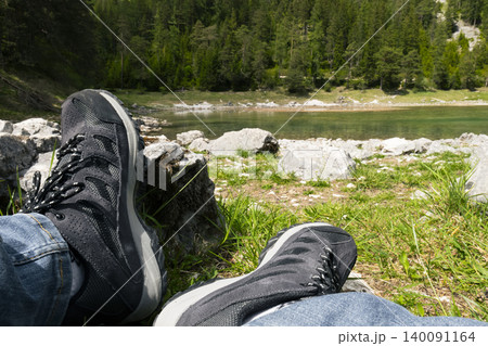 Person in hiking boots relaxing by a lake surrounded by forest, highlighting an adventure lifestyle and connection with nature. Person in hiking boots relaxing by a lake surrounded by forest, highlighting an adventure lifestyle and connection with nature. 140091164