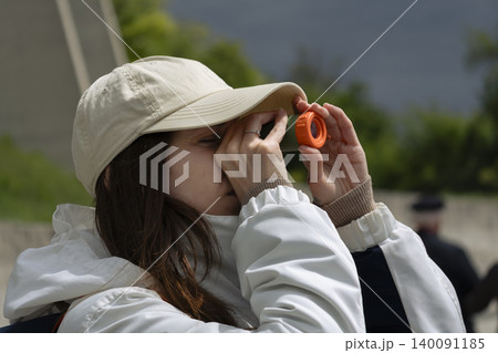 Young woman looking through a small orange telescope outdoors, showcasing a portable magnification device for everyday use. 140091185