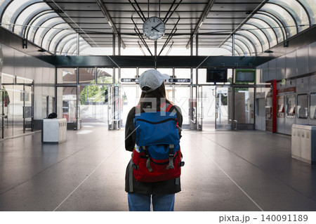 Young woman with a backpack standing at the train station beside a large round clock, symbolizing accurate timekeeping. 140091189