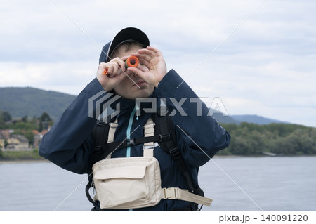 Man looking through a small orange telescope at a river landscape, showcasing a portable magnification device for everyday use. 140091220