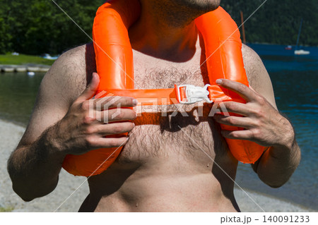 Man wearing an orange inflatable life vest standing on a lake shore, highlighting water safety during outdoor recreation Man wearing an orange inflatable life vest standing on a lake shore, highlighting water safety during outdoor recreation 140091233