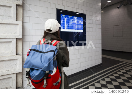 Young woman with a backpack looking at the digital departure board at a train station, checking the train timetable. 140091249