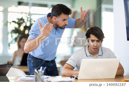 Man scolding sad male colleague working on laptop in office 140092287