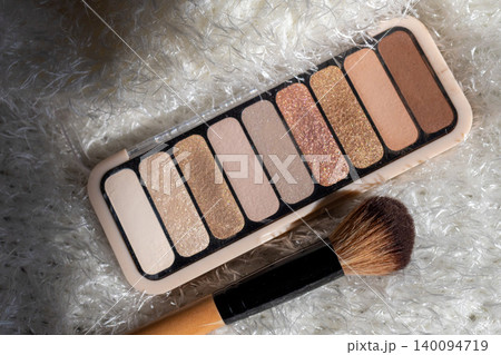 Eyeshadow palette displaying a range of neutral nude and glitter shades for everyday makeup looks, next to an applicator brush on a soft white furry background Eyeshadow palette displaying a range of neutral nude and glitter shades for everyday makeup looks, next to an applicator brush on a soft white furry background 140094719
