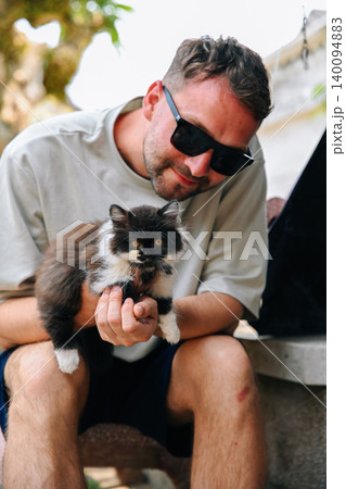 A caucasian 30 years old young man sitting outdoors and holding a cute fluffy domestic kitten in hands A caucasian 30 years old young man sitting outdoors and holding a cute fluffy domestic kitten in hands 140094883