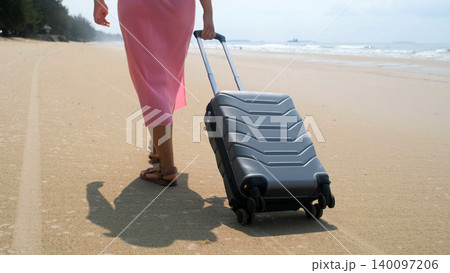 Close rear view of young woman in pink dress rolling a grey suitcase along sandy beach with sea waves nearby. Summer vacation concept 140097206