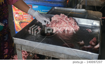 Close-up of woman's hands pushing fresh meat pieces into a large electric meat grinder at a covered Thai market stall. Mincing process concept 140097207