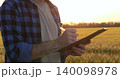 Close-up of male farmer holding pen and clipboard, writing notes during crop inspection in wheat field at sunset. Concept of agricultural documentation, crop control, and farm work. 140098978