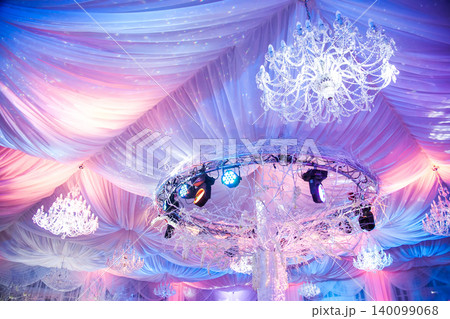 Luxurious event ceiling with crystal chandeliers, draped fabric, and pink and purple lighting Luxurious event ceiling with crystal chandeliers, draped fabric, and pink and purple lighting 140099068