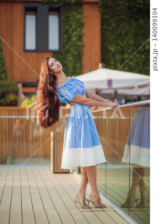 Stylish young woman with long brown hair in blue dress on modern balcony 140099104
