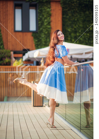 Joyful woman with long hair in blue dress posing playfully on modern terrace 140099105