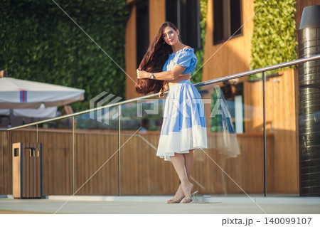 Elegant woman with long brown hair in blue summer dress poses on modern terrace Elegant woman with long brown hair in blue summer dress poses on modern terrace 140099107