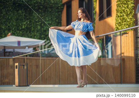 Elegant young woman in blue summer dress twirling skirt on modern sunny terrace Elegant young woman in blue summer dress twirling skirt on modern sunny terrace 140099110