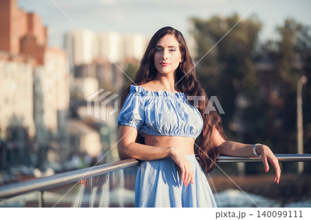 Confident young woman with long dark hair in blue dress posing on city balcony during sunset Confident young woman with long dark hair in blue dress posing on city balcony during sunset 140099111