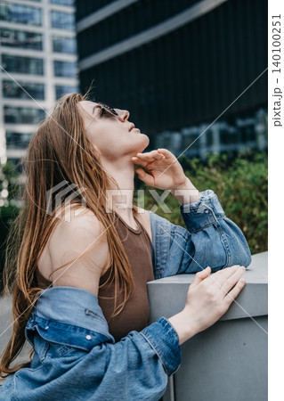 Woman wearing sunglasses tilts head up while leaning against outdoor structure in corporate district. Stress management metropolitan, taking a mindful break, personal resilience city dweller. Woman wearing sunglasses tilts head up while leaning against outdoor structure in corporate district. Stress management metropolitan, taking a mindful break, personal resilience city dweller. 140100251