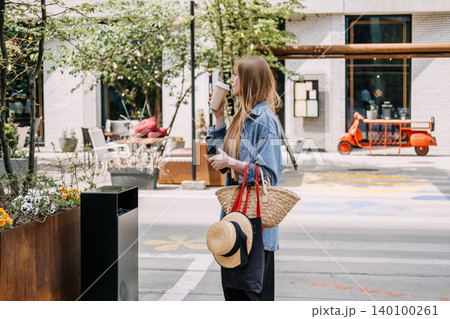 Young woman drinks from cup while holding smartphone on urban street. Zero waste daily routine, sustainable textile accessories, conscious retail choices. Young woman drinks from cup while holding smartphone on urban street. Zero waste daily routine, sustainable textile accessories, conscious retail choices. 140100261