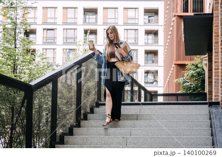 Woman walks down outdoor city staircase while holding coffee cup and smartphone. Freelancer on the move, digital nomad daily routine, remote work lifestyle. 140100269