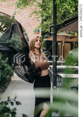 Woman holds up sheer black fabric while posing on outdoor cafe patio. Urban fashion portrait, contemporary street style, modern female look. 140100271