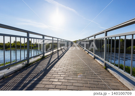 Sunlit pedestrian bridge crossing a calm river, featuring a paved walkway 140100709
