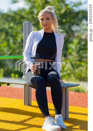 Woman resting outdoors in activewear holding water bottle on a sunny day 140103429