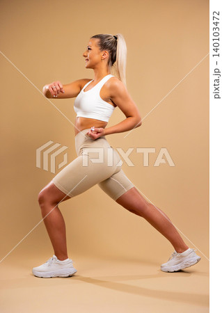 Athletic woman in white sports bra and beige leggings performing lunges in studio setting Athletic woman in white sports bra and beige leggings performing lunges in studio setting 140103472