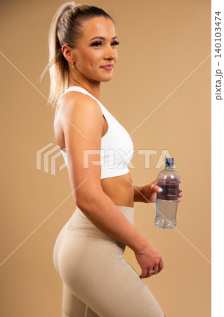 Fit woman in white sports bra and beige leggings holding water bottle standing in studio 140103474