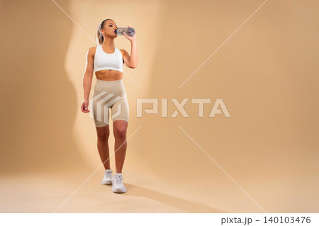 Fit athletic woman in white sports bra and beige leggings drinking water from bottle during workout session 140103476