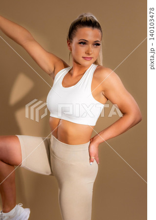 Fit young woman in white sports bra and beige leggings posing confidently in fitness wear Fit young woman in white sports bra and beige leggings posing confidently in fitness wear 140103478