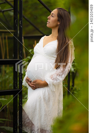 Pregnant woman in white dress reflecting in a lush green garden 140103489