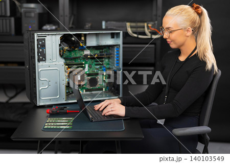 Female IT technician in her twenties works on laptop beside open server for diagnostics Female IT technician in her twenties works on laptop beside open server for diagnostics 140103549