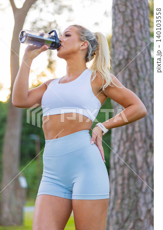 Woman in activewear drinking water outdoors after a fitness routine Woman in activewear drinking water outdoors after a fitness routine 140103558
