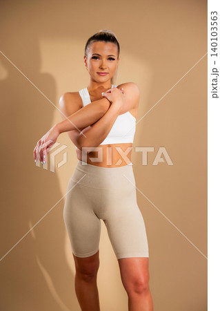 Fit woman in white sports bra and beige ribbed leggings stretching in studio Fit woman in white sports bra and beige ribbed leggings stretching in studio 140103563