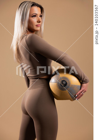 Fit woman in brown workout gear holding a gold medicine ball in studio setting Fit woman in brown workout gear holding a gold medicine ball in studio setting 140103567
