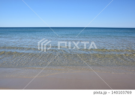 A serene beach landscape featuring clear blue water meeting a soft sandy shore, with gentle waves creating a calming effect under a clear sky, ideal for relaxation and leisure 140103769
