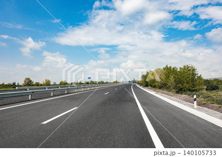 Wide highway with clear blue sky, guardrail, roadside vegetation 140105733