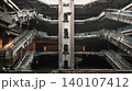 The skeletal ruins of an abandoned shopping mall rise symmetrically around a central shaft, their rusted escalators frozen over a dark, flooded floor. Patches of wild green foliage drape softly across 140107412