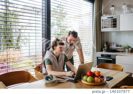 Father helping teenage son with diabetes with homework on laptop in kitchen. Father helping teenage son with diabetes with homework on laptop in kitchen. 140107977