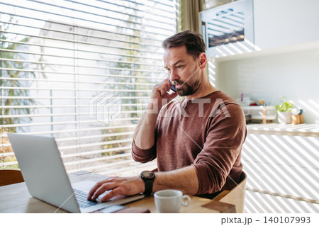 Man sitting in kitchen working remotely on laptop and making phone call. Man sitting in kitchen working remotely on laptop and making phone call. 140107993