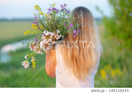 Woman carrying a large bouquet of wildflowers in a lush green field during sunset Woman carrying a large bouquet of wildflowers in a lush green field during sunset 140110243