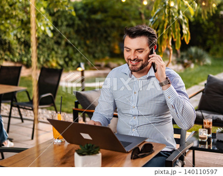 Smiling man using laptop and talking on phone at outdoor cafe Smiling man using laptop and talking on phone at outdoor cafe 140110597