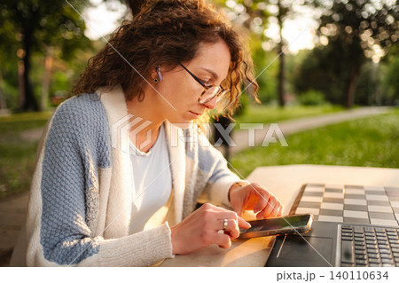 Young woman working remotely with smartphone and laptop in park at sunset 140110634