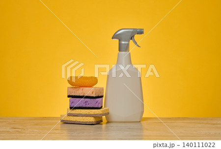 A white plastic spray bottle and a stack of multi-colored sponges stand on a wooden surface 140111022