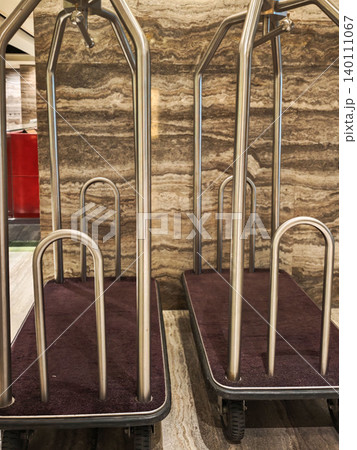 Hotel luggage carts with metal frames and plush fabric bases positioned against a textured stone wall in a modern lobby area Hotel luggage carts with metal frames and plush fabric bases positioned against a textured stone wall in a modern lobby area 140111067