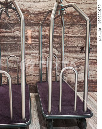 Two metal luggage carts with purple fabric bases positioned against a textured stone wall in a hotel lobby area Two metal luggage carts with purple fabric bases positioned against a textured stone wall in a hotel lobby area 140111070