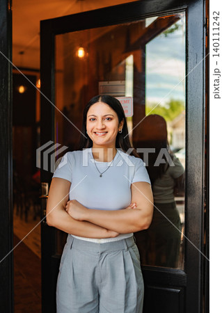 Smiling woman standing with arms crossed at entrance of coffee shop Smiling woman standing with arms crossed at entrance of coffee shop 140111242